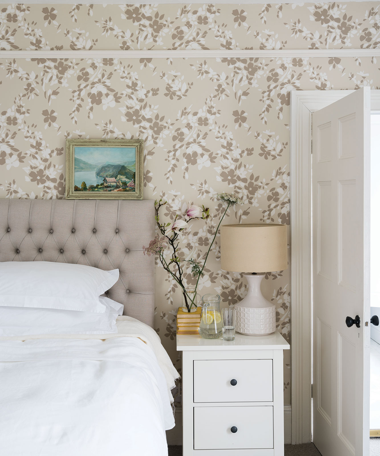 Floral patterned bedroom wallpaper ideas in beige, with white bedroom furniture and a small gold-framed painting resting on the headboard.