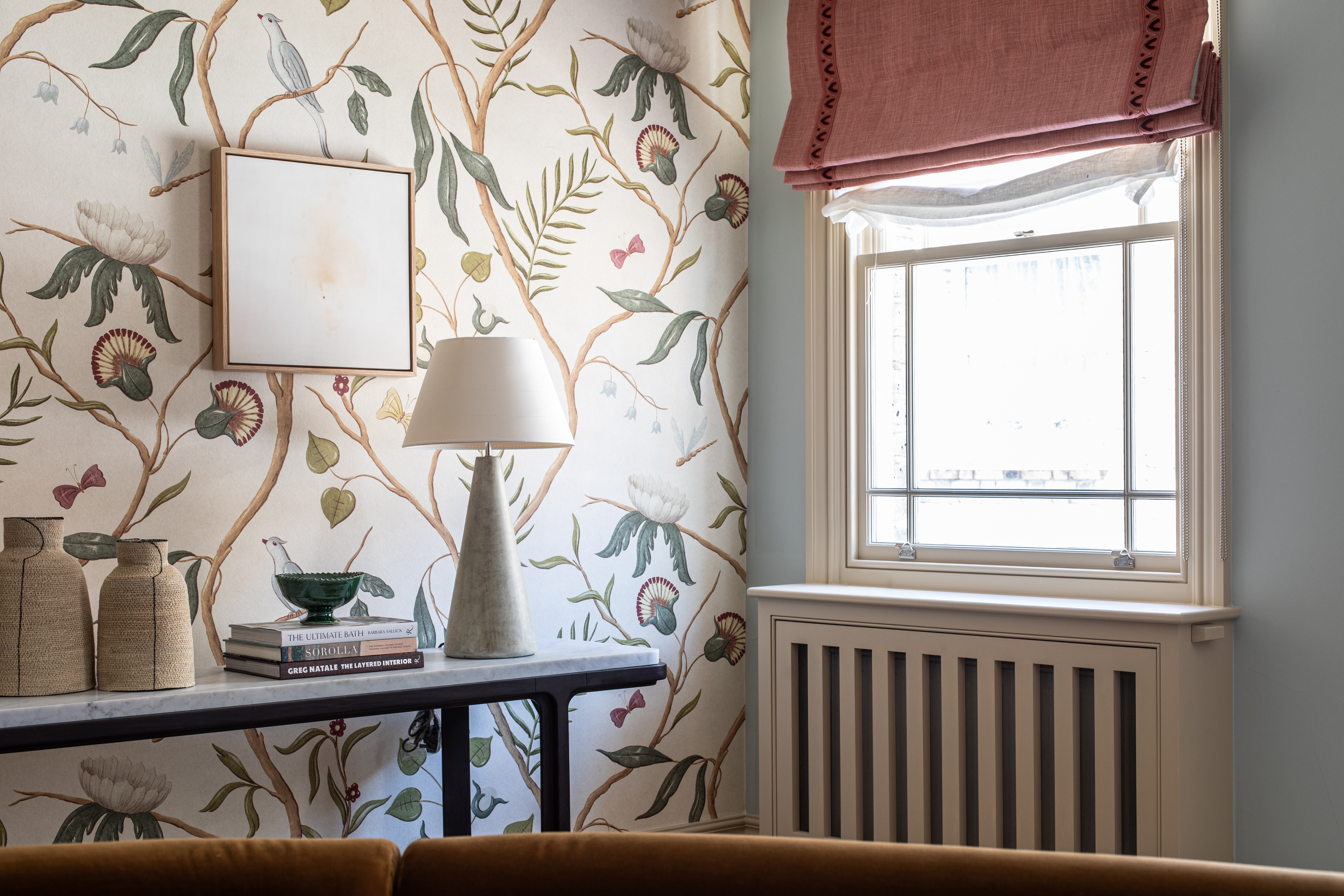 a living room corner with a radiator cover below the window with a dusty pink roman blind, a table with a lamp and decorative objects sits in front of a wall with ornate wallpaper
