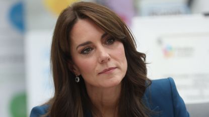 Kate Middleton has long brunette hair and side swept bangs and is wearing a dark blue suit jacket