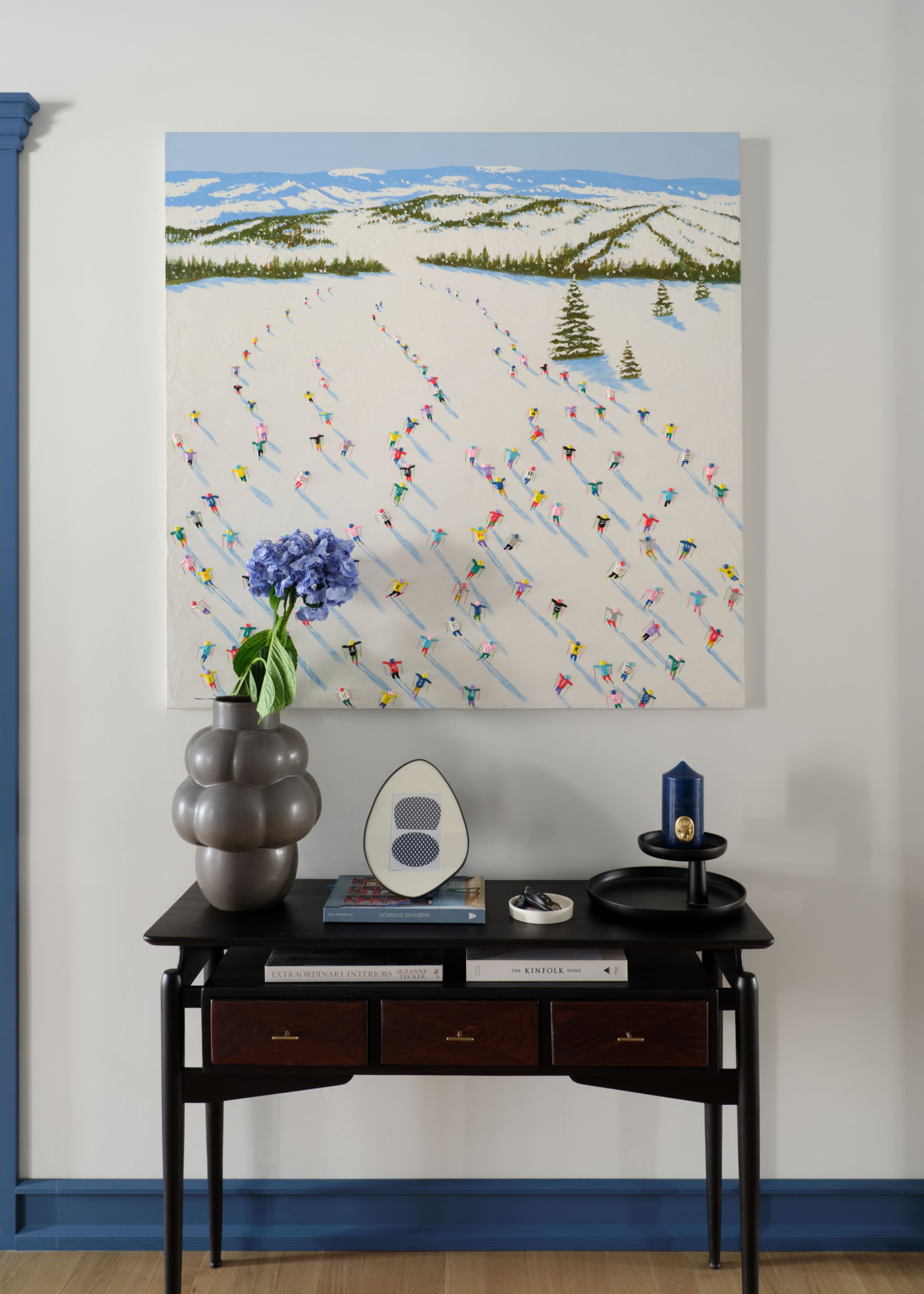 A painting of skiers above a black console table with a bubble-like flower vase