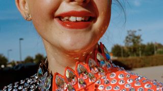 A tight crop of a smiling young dancer's face and throat, her red lips and gap-toothed grin set above a spectacular orange costume collar encrusted with large iridescent crystal gems.