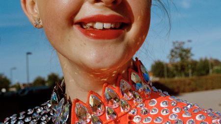 A tight crop of a smiling young dancer's face and throat, her red lips and gap-toothed grin set above a spectacular orange costume collar encrusted with large iridescent crystal gems.