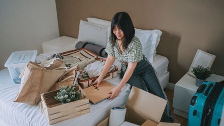 A woman sat on her bed decluttering her bedroom, tidying away boxes and items as well as suitcases
