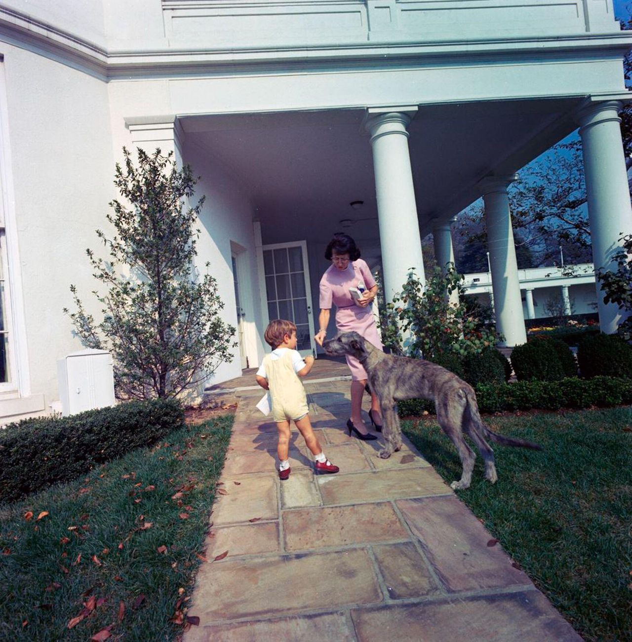 A colour photograph of a young John F. Kennedy Jr. and Evelyn Lincoln, President Kennedy&rsquo;s secretary, interacting with an Irish Wolfhound named Wolf. They are standing on a stone path outside the Oval Office, with the bright white columns of the West Wing and neatly kept garden visible in the background. It is a sunny day, and the scene is relaxed and informal.