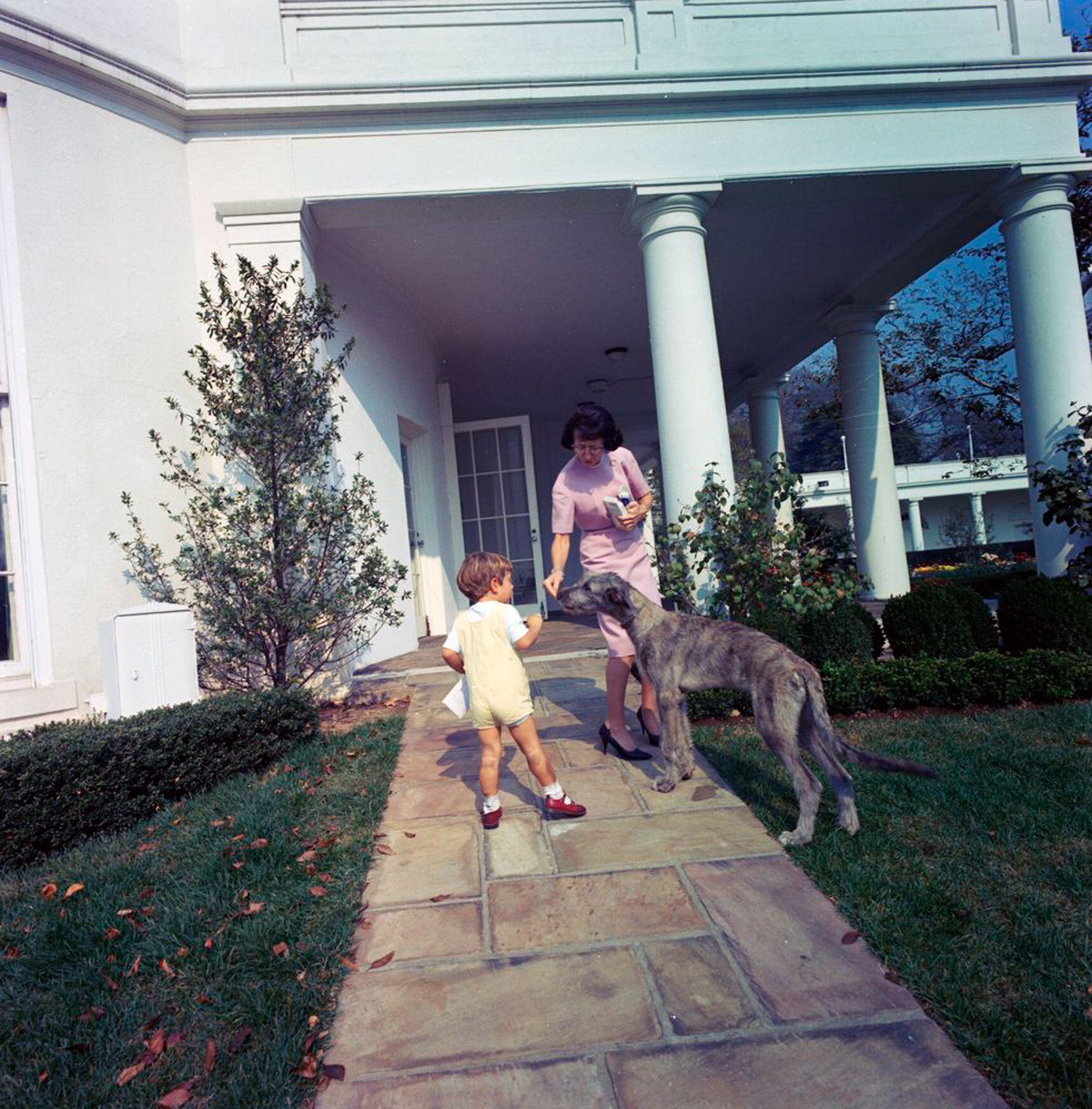 A colour photograph of a young John F. Kennedy Jr. and Evelyn Lincoln, President Kennedy’s secretary, interacting with an Irish Wolfhound named Wolf. They are standing on a stone path outside the Oval Office, with the bright white columns of the West Wing and neatly kept garden visible in the background. It is a sunny day, and the scene is relaxed and informal.