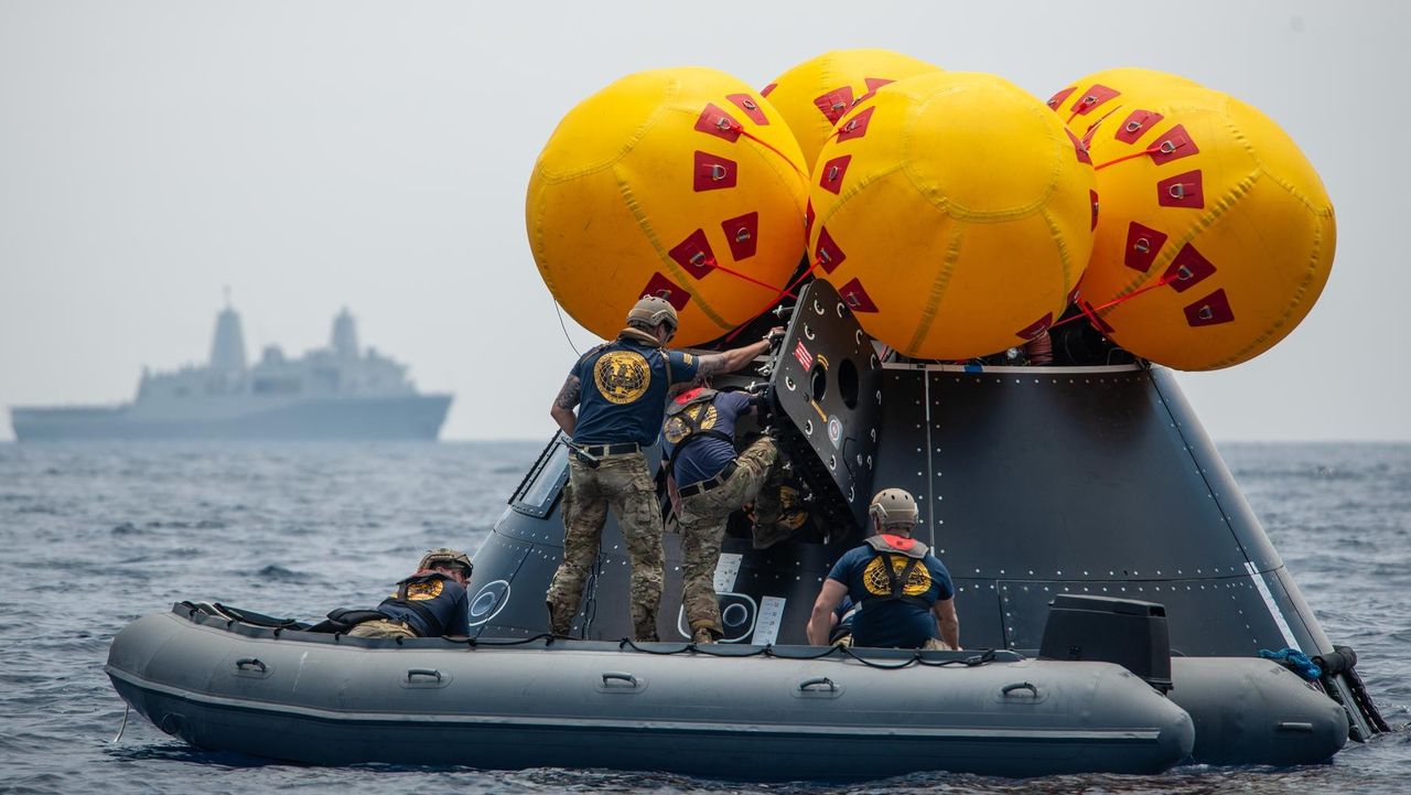 U.S. Navy divers enter a simulated Artemis 2 spacecraft, called the Orion Crew Module Test Article, during practice recovery operations on July 28, 2023. Here they are pretending to recover astronauts and lift them on to an inflatable "front porch" for extraction to a waiting helicopter. Their destination, the U.S.S. John P. Murtha warship, is visible in the background.