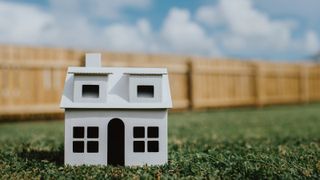 Simple image of a plain, white, cardboard house sitting on grass, with a fence in the background. Cloudy blue sky provides a space for copy.