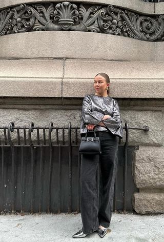 A woman standing outside wearing gold earrings, a silver jacket, black jeans, silver pumps, and a black handbag.