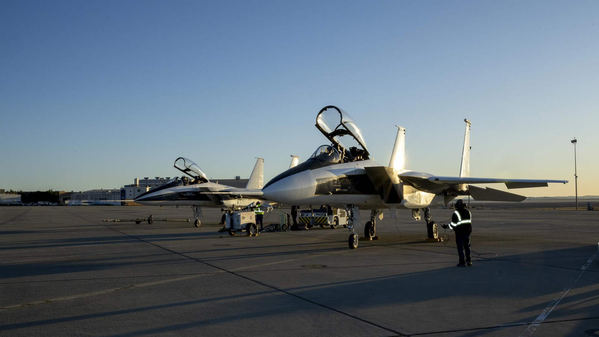 Two large gray planes with their cockpits open sit on a tarmac with a sunset behind them
