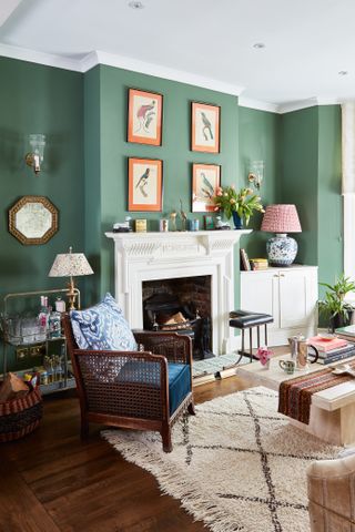 A classic traditional living room with green wall decor in London home with cream colored fireplace, lampshade decor, framed wall art decor, cream tasselled rug decor and wall lighting