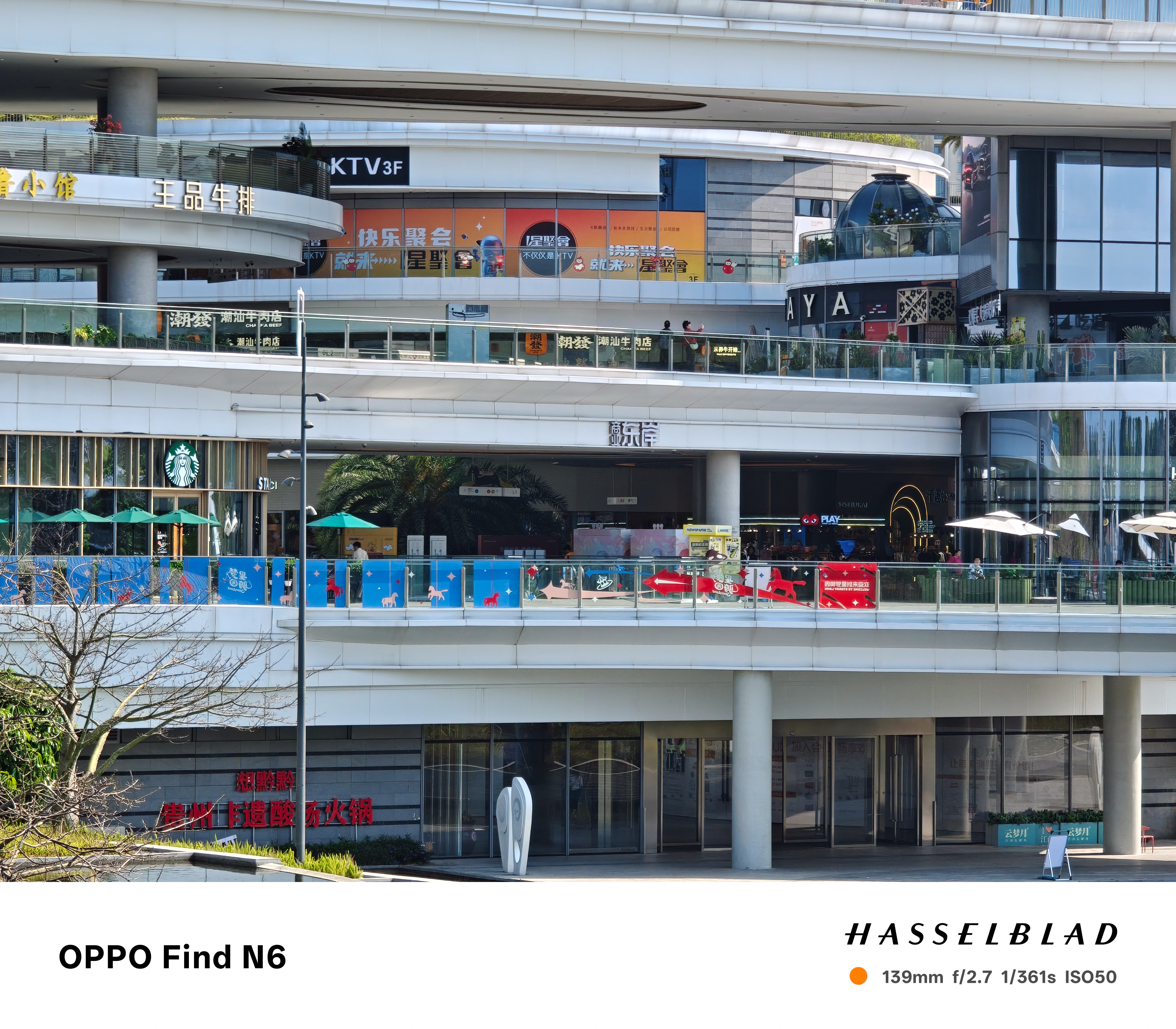 A close-up telephoto photograph of a specific section of the shopping complex, focusing on a Starbucks storefront and a walkway decorated with blue and red banners. The image demonstrates high clarity on the distant text and architectural details.