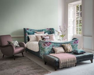 A feminine bedroom with teal floral patterned bed, dusty pink upholstered accent chair and ottoman at end of bed