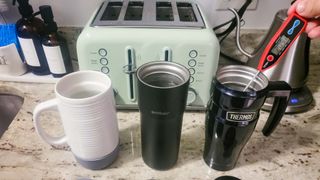 Best travel coffee mugs: Ello Jane Ceramic Travel Mug, Ember and Thermos shown on counter with digital thermometer testing liquid temperature.