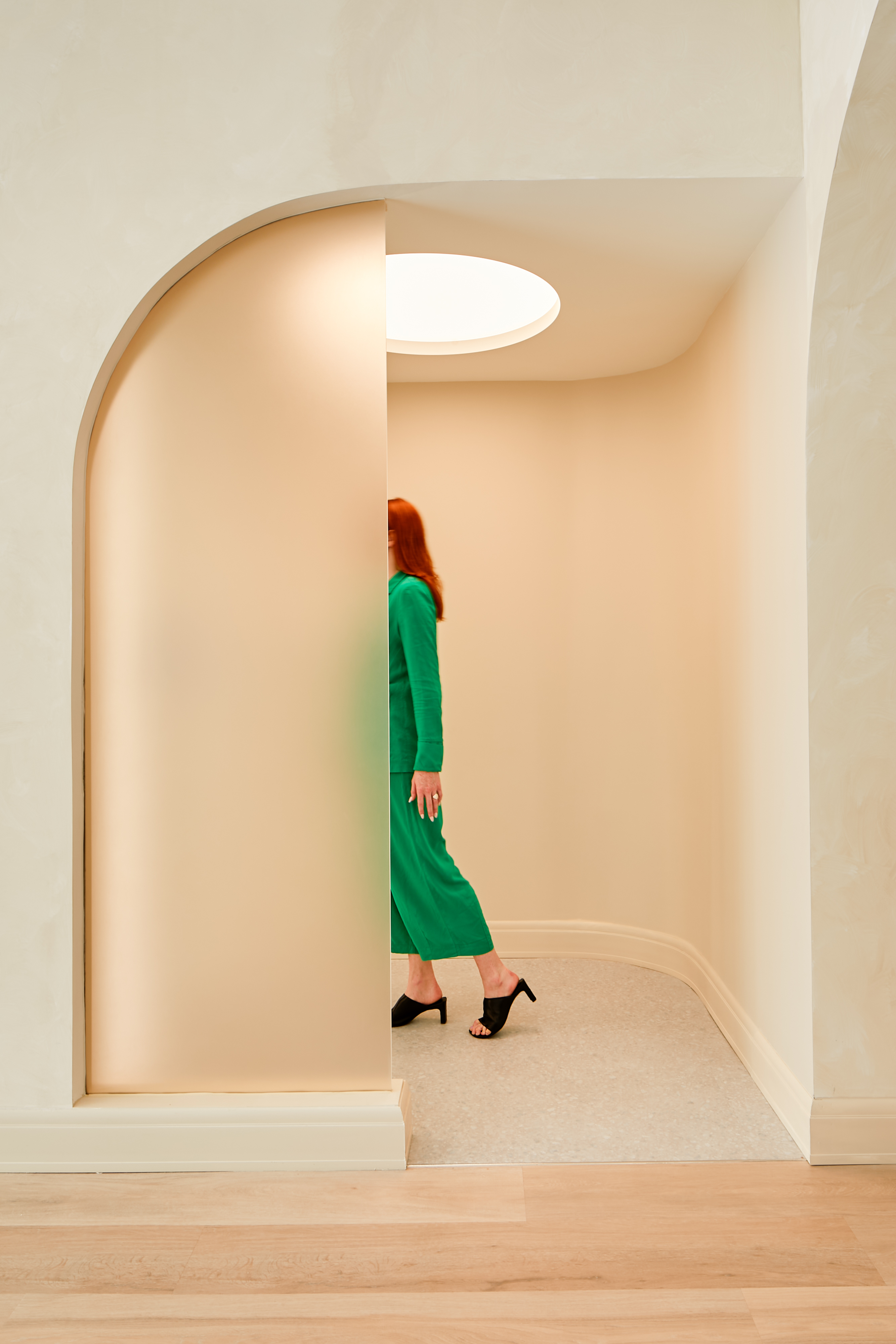 A woman dressed in an emerald green maxi dress and black, open-toe mules is captured as she walks into a corridor positioned pasted an arched, matte glass-decorated doorless passageway.