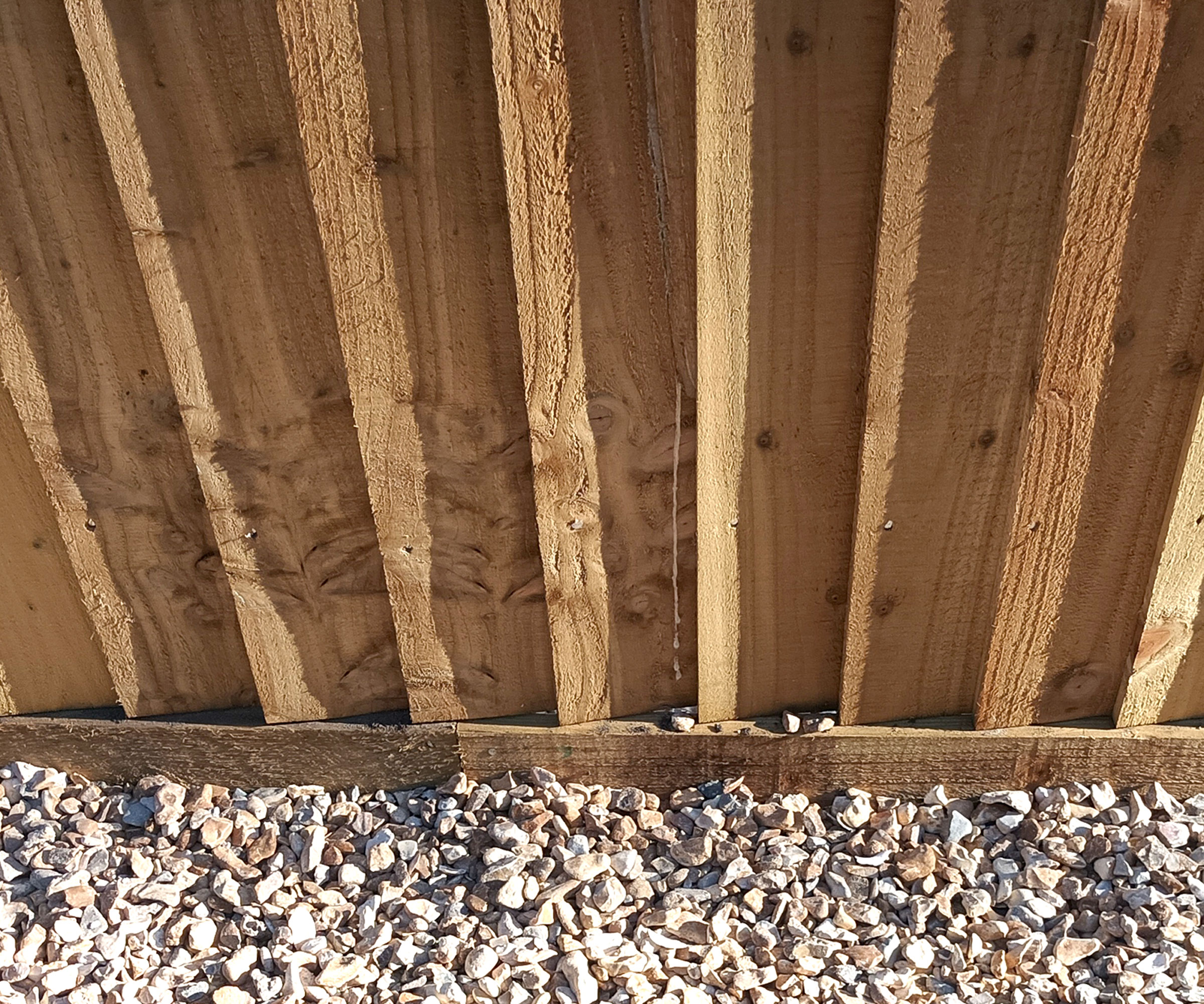 Close up of bottom of feather edge fencing showing barge board against light coloured stones