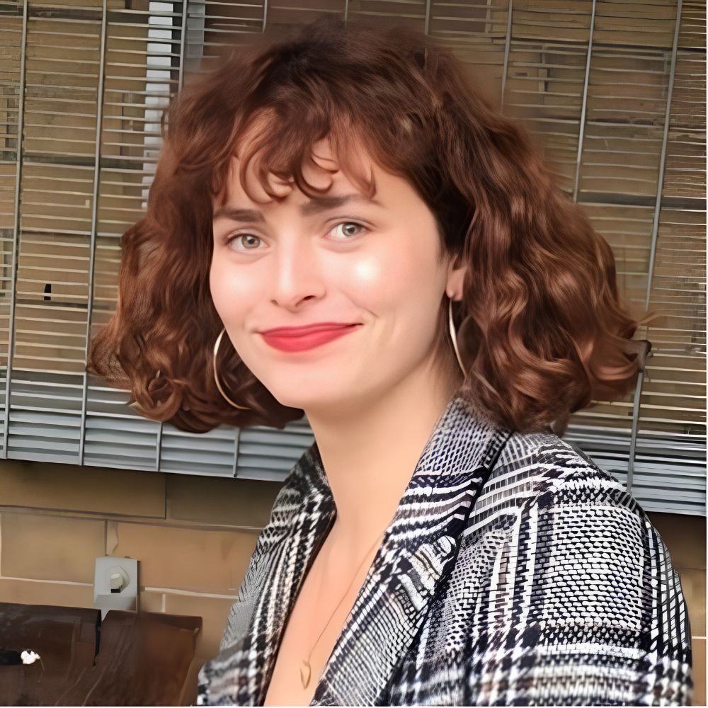 A smiling woman with short curly auburn hair and a fringe, wearing gold hoop earrings, red lipstick and a grey checked blazer.