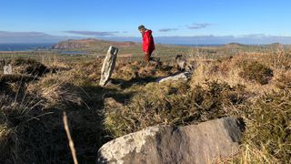 Archaeologist in red coat stands on grassy seaside cliff before a large rock marking a burial site.