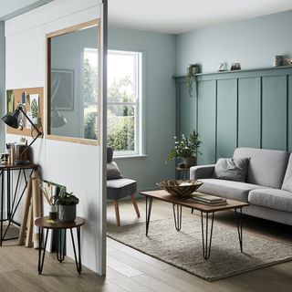 a living room with wooden floors divided by a room divider with a window, one side of the living room visible with a green panelled feature wall in front of a grey sofa