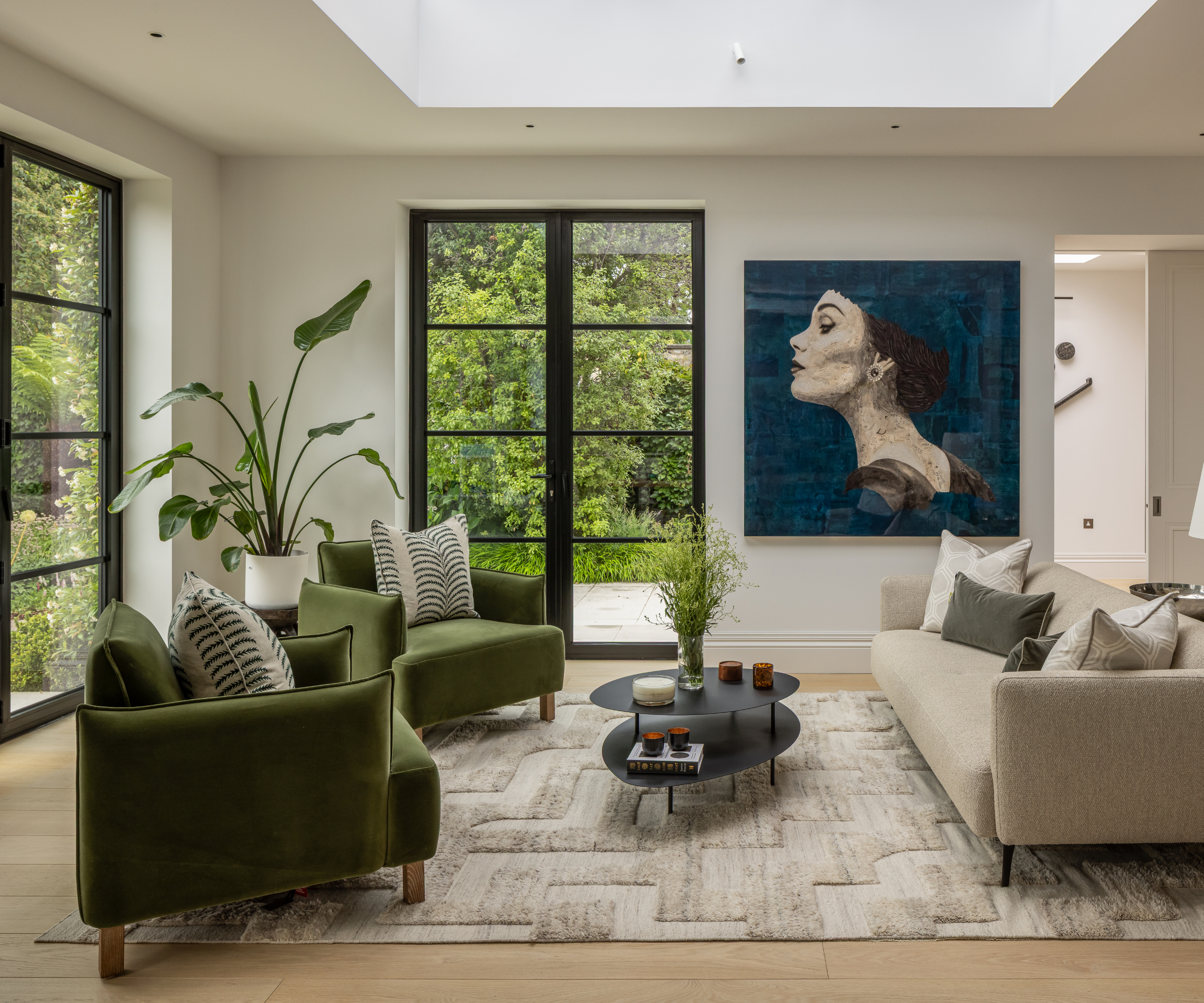 living room with large rooflight, two set of black crittall doors, green and cream sofas, wooden floor and large rug, bold portrait canvas on wall