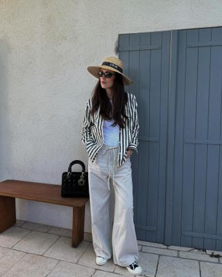 Woman wears a Dior straw hat, drawstring linen trousers, black canvas sneakers and a black and white striped button up.