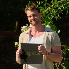 reality star harry jowsey stands in front of a floral display holding a sign that says my vows in a still from the show let's marry harry