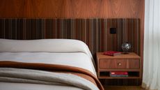 modern bedroom with white bedcover, with rust and beige throws folded over the end of the bed. the wall behind the bed has a timber veneer and an upholstered headboard with floating timber side tables