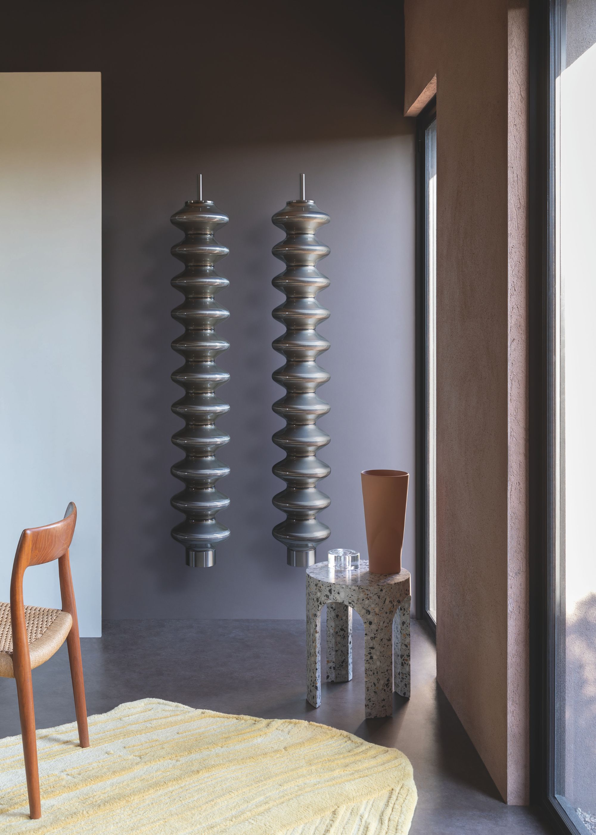 two wall-mounted grey radiators in a home. Beside the radiator is a table with a glass and a oak chair sits on the side.