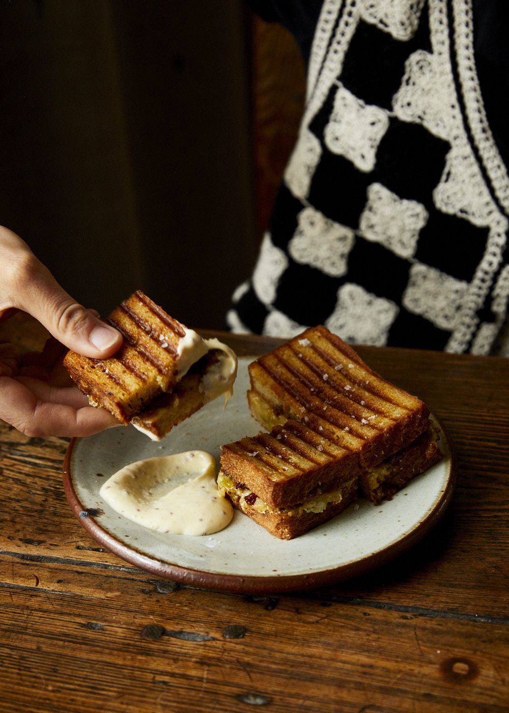 A cheese toasties sliced into four is being dipped into a mayo and mustard dressing by someone wearing a checkered white and black vest.