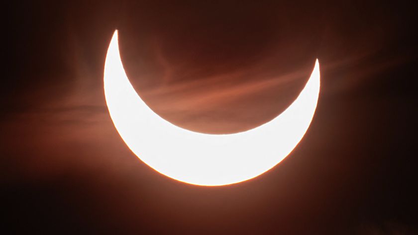 The sun&#039;s disk appears as a glowing upturned crescent shining against a dark sky framed by wispy clouds during a partial solar eclipse.