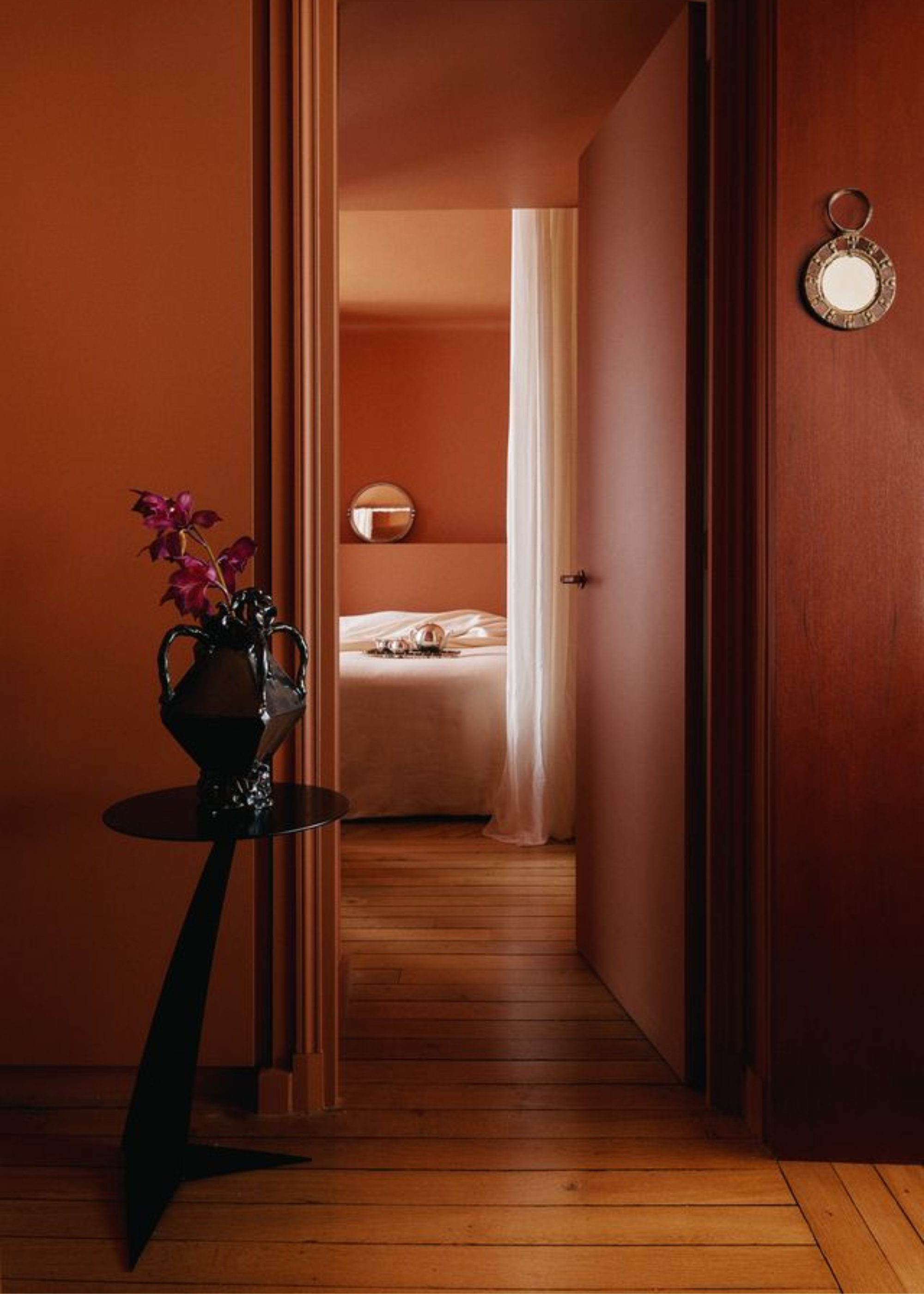 A doorway leading to a bedroom with a pale pink bedscape, and a tray with a silver tea set, a small circular mirror, and a pedestal with a vase of flowers