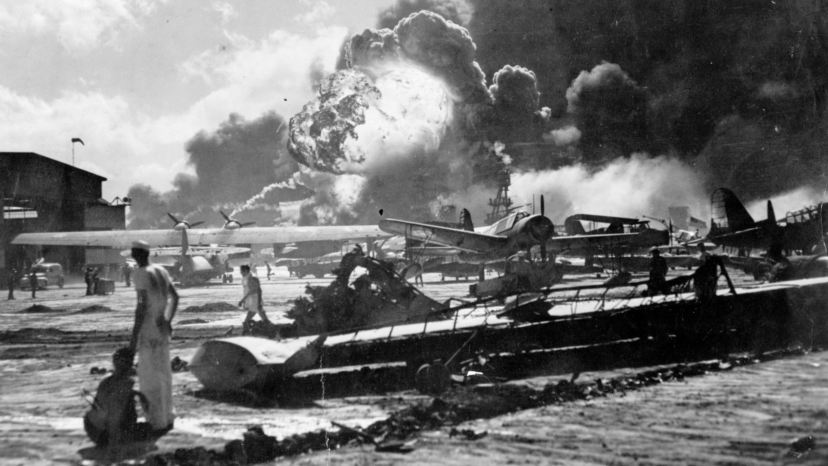 During the attack on Pearl Harbor, an explosion is seen at the Naval Air Station, Ford Island, on the southern coast of Oahu. Sailors stand amid wreckage watching as the USS Shaw explodes in the center background. The USS Nevada is also visible in the middle background, with her bow headed toward the left.