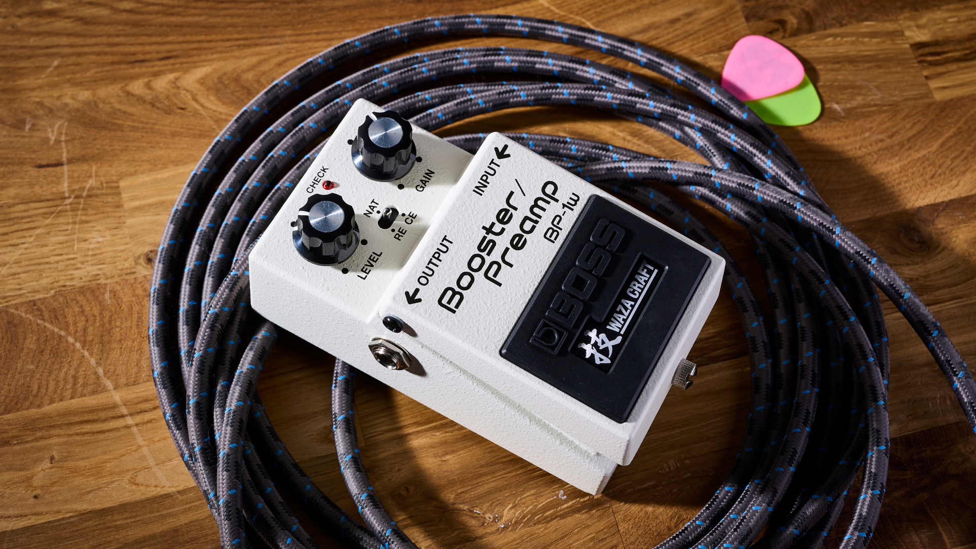 Boss Waza Craft BP-1W Boost/Preamp: two classic preamps in one compact, white stompbox, here pictured with green and pink guitar picks, and a guitar cable coiled around it.