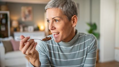 close up shot of a woman wearing a striped white and grey top looking sideways to the camera holding a spoon of food to her mouth. she has short grey hair and there's a living room setting behind her.
