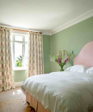 A light green bedroom with a window with long white purple flower patterned drapes, a bed with a pink headboard and white bedding with white and purple sprigs next to it, and a scalloped jute rug at the foot of it