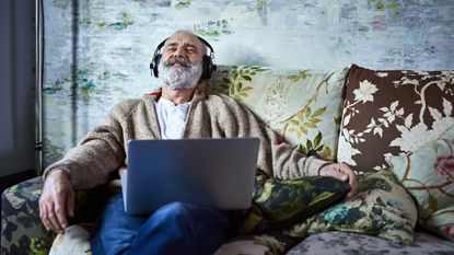 An older guying wearing headphones relaxes on his sofa.