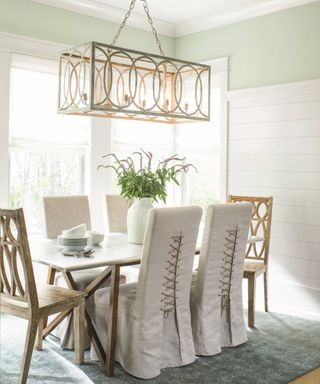 White and green dining room with a hint of pale green, large chandelier, vintage style chairs, rug, table and vase,