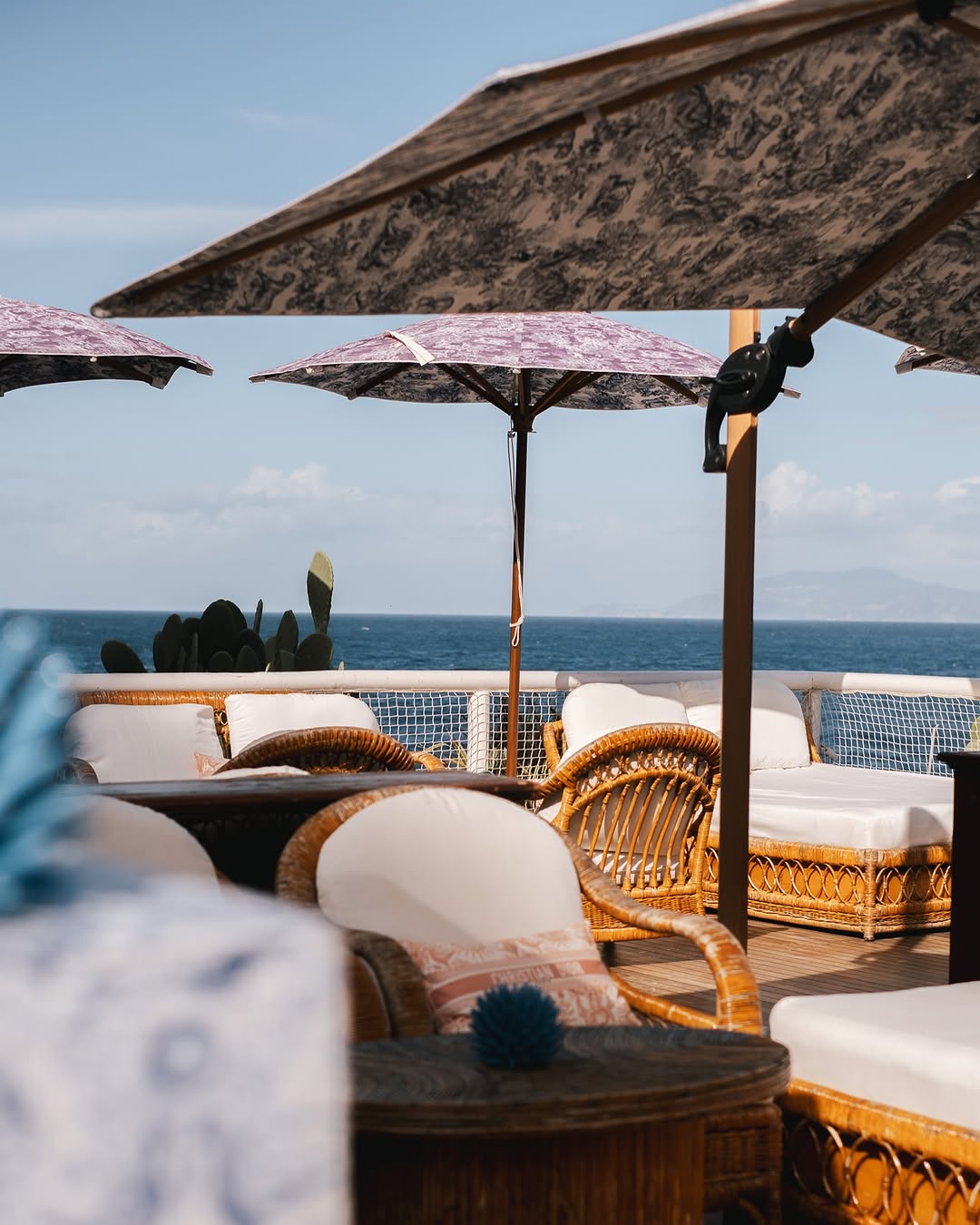 A sun-soaked terrace with rattan and textile loungers and floral parasols in blue and white fabric and wood.