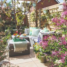 A blooming garden with a corner outdoor sofa in grey rattan effect with grey seat cushions and blue-green outdoor scatter cushions