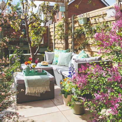 A blooming garden with a corner outdoor sofa in grey rattan effect with grey seat cushions and blue-green outdoor scatter cushions