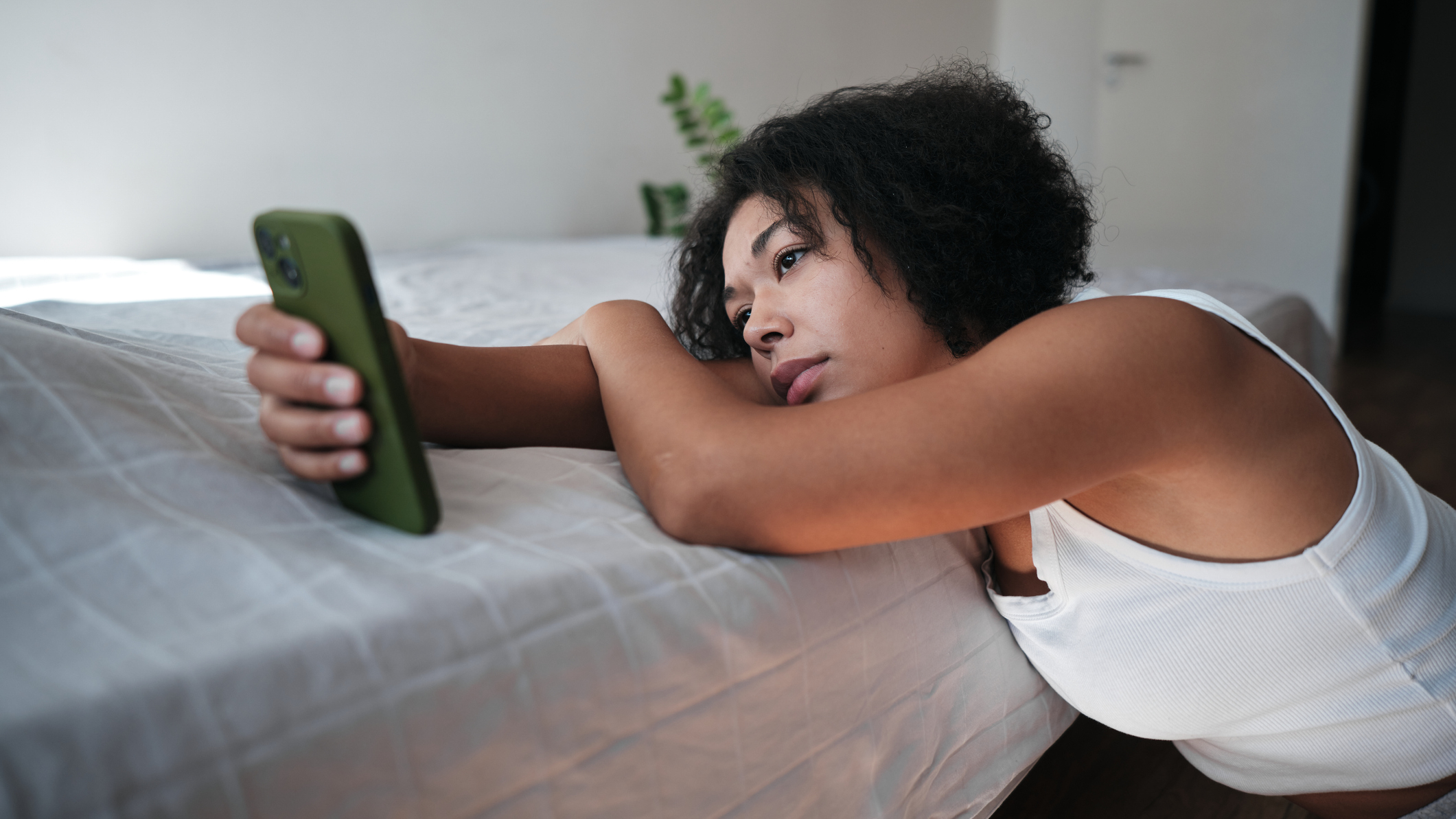 Woman rests her head on the edge of a bed and stares passively at a smartphone
