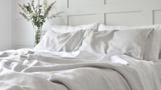 A bed with slubby white linen bedding on it. A glass vase of eucalyptus leaves in the background. A white painted panelled wall behind.