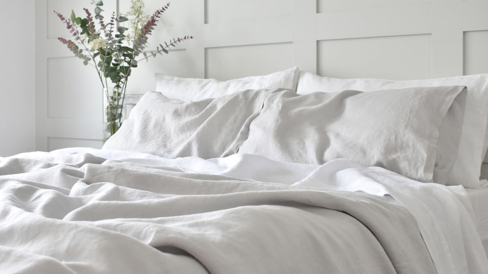 A bed with slubby white linen bedding on it. A glass vase of eucalyptus leaves in the background. A white painted panelled wall behind. 