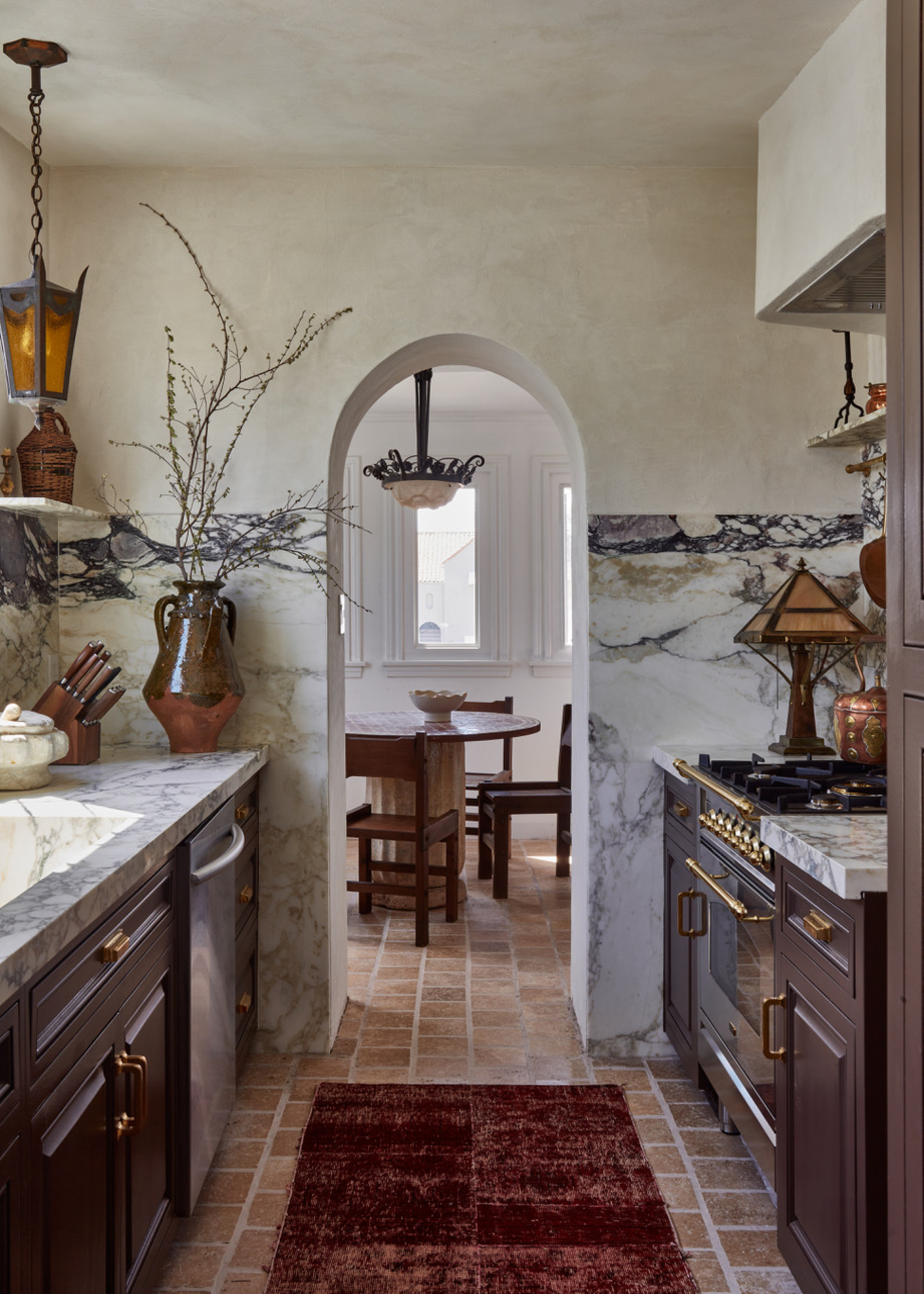 A small galley kitchen with burgundy cabinets, terracotta floors, and high-contrast marble countertops