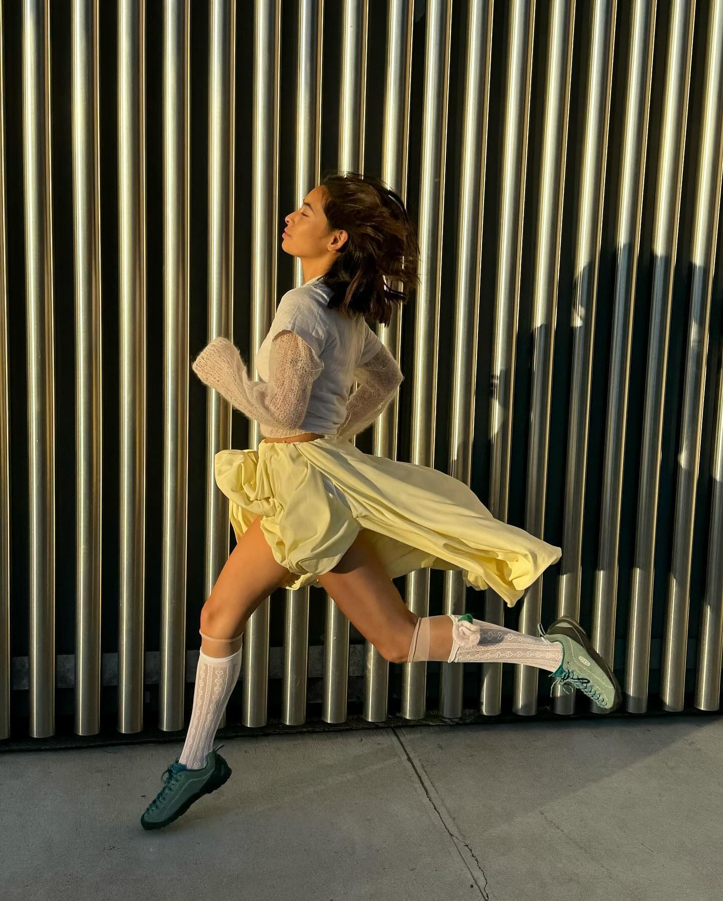Influencer @himichelleli jumps through the air wearing green Keen trainers with a white skirt and grey top.