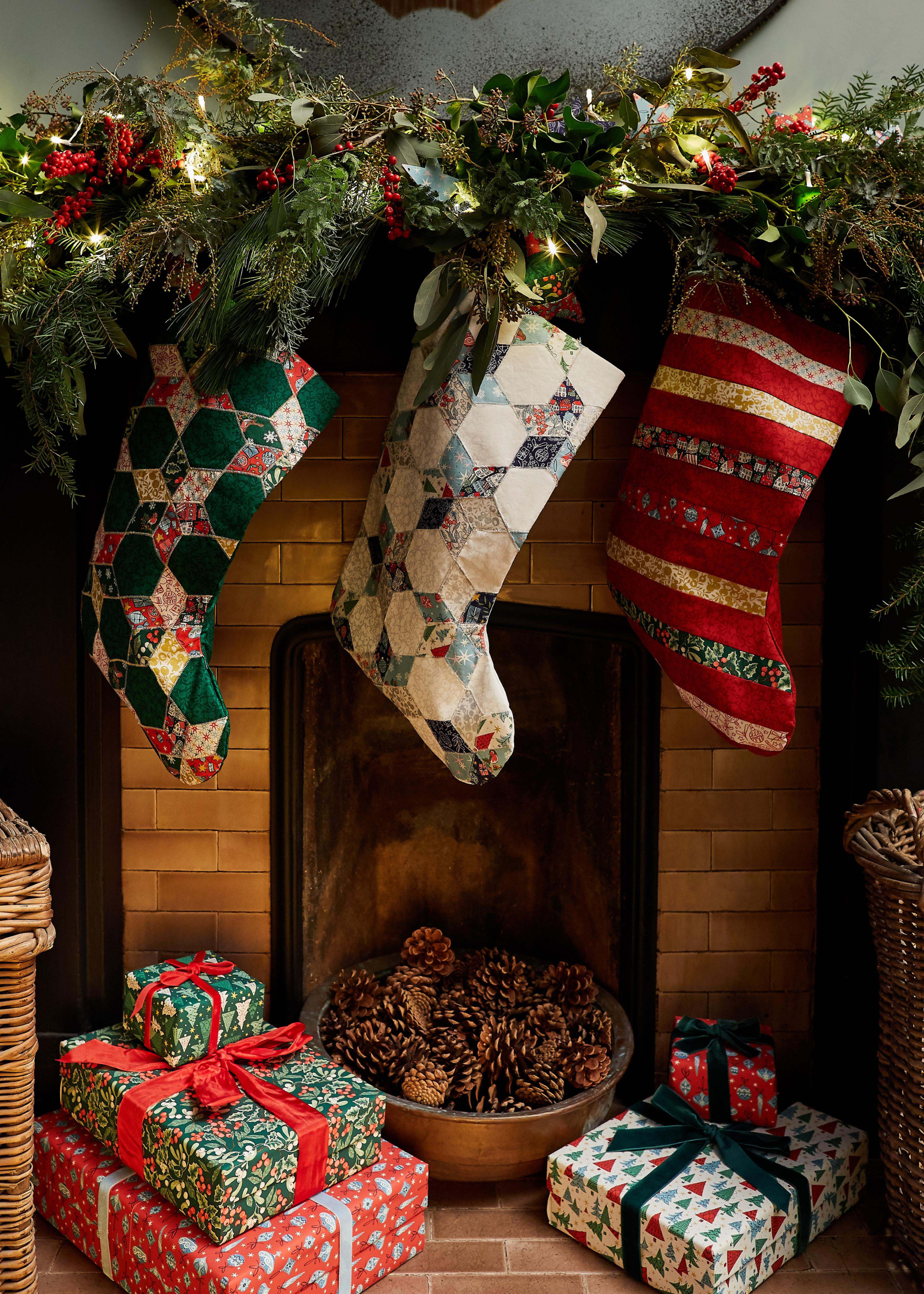 A Christmas fireplace with three stockings in Liberty prints hung from the mantel with a lit garland over the top and presents underneath and either side a large of bowl of pinecones inset into the empty fireplace