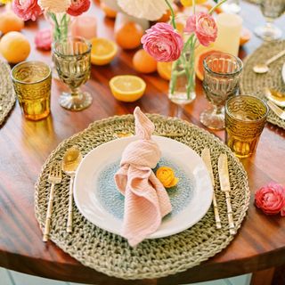 Summer tablescape with woven placemat, pink napkin, coloured glassware, flowers and fresh oranges