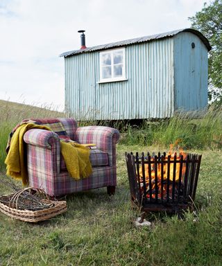 A pale blue shepherd's hut in a meadow with a tartan armchair and fire pit.