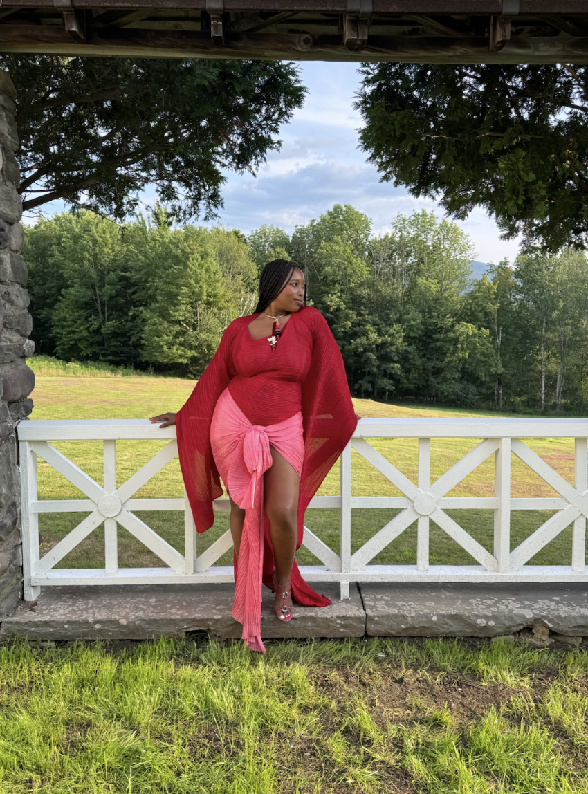 Mecca James-Williams in a pink and red wedding guest dress leaning against a white fence