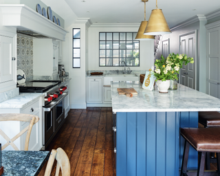 A blue kitchen island with a white and gray granite worktop with dark wood flooring and two gold pendant lamps