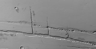 A grayscale scene with features on the surface of this world, which is viewed from an above angle. Some look like a dotted path and other look like long streaks.
